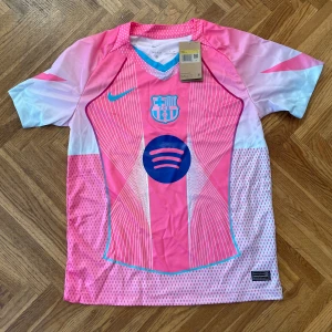 Barça x Nike rosa fotbollströja S - Snygg FC Barcelona fotbollströja från Nike i rosa och vitt med blå detaljer. Tröjan har korta ärmar, V-ringning med turkos kant och coolt grafiskt mönster framtill. Materialet är lätt och andas, Dri-Fit. Spotify-logga och klubbmärke på bröstet.