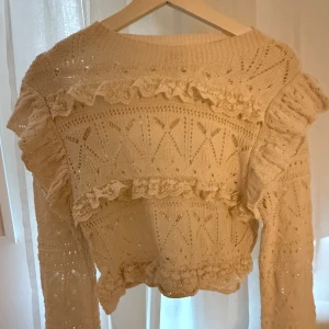 Beige stickad tröja med volanger - Superfin beige stickad tröja med volangdetaljer och snyggt hålmönster. Tröjan är croppad och har långärmade ärmar.