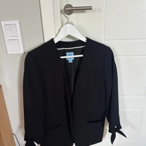 Svart kavaj från CeCe med knyt - Stilren svart kavaj från CeCe med snygga knytband vid ärmsluten. Klassisk rak passform och två fickor framtill. Perfekt för dig som vill ha en enkel men trendig look. Passar till allt och är lätt att matcha med andra plagg.