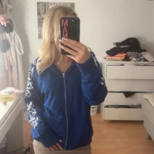 Blå zip-hoodie med tryck på ärmar - Blå hoodie med dragkedja och huva, samt vita tryck längs båda ärmarna i coolt typsnitt. Jackan har fickor framtill och en relaxed passform. Perfekt för dig som gillar streetstyle och vill ha något bekvämt och snyggt.