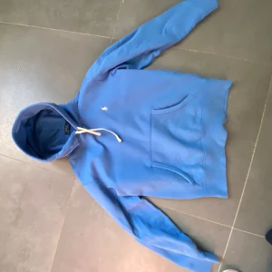 Blå hoodie från Polo Ralph Lauren - Säljer en chill blå hoodie från Polo Ralph Lauren i storlek S. Tröjan har vit snörning vid huvan, klassisk känguruficka och den ikoniska Polo-loggan broderad på bröstet. Perfekt för dig som gillar stilren och bekväm streetwear.