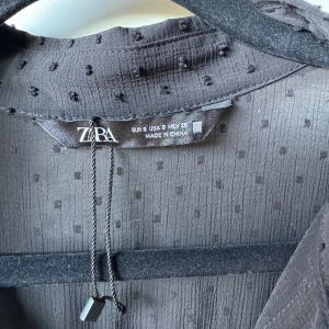 Svart prickig klänning från Zara - Supersnygg svart klänning från Zara med diskret prickigt mönster. Klänningen har v-ringning, knappar framtill och smock i midjan som ger en cool siluett. Långa ärmar med manschett och tillverkad i lätt, transparent polyester. Perfekt för dig som gillar stilrena detaljer.