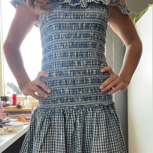 Blåvit rutig kortklänning med volang - Supersöt blåvit kortklänning med rutigt mönster och smockad överdel. Klänningen har offshoulder-ärmar med volangdetaljer upptill och en luftig kjol. Perfekt för varma dagar och ger en fräsch och ungdomlig vibe.