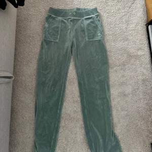 Juicy  couture  - Snygga gröna mjukisbyxor i mjukt velourmaterial med bred resår i midjan och stora fickor fram. Baktill finns två fickor med knappar. Perfekta för chill dagar och har en relaxed passform. Färgen är dämpad grön och byxorna har raka ben.