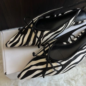 Zebramönstrade ballerinaskor från NA-KD - Snygga slingback flats från NA-KD med zebramönster i svart och vitt. Skorna har spetsig tå, rosett framtill och öppen häl. Aldrig använda därför det säljes, skickas i orginallåda. Perfekt för dig som vill sticka ut med coola detaljer. Storlek 38. 