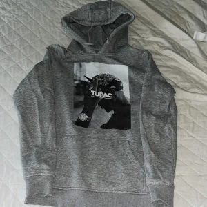 Grå Tupac hoodie från Mister Tee - Säljer en grå hoodie från Mister Tee med stor svartvit Tupac-print på framsidan. Tröjan har huva med snörning, magficka och långa ärmar. Perfekt för dig som gillar streetstyle och vill ha en chill look med ikonisk hiphop-vibe.