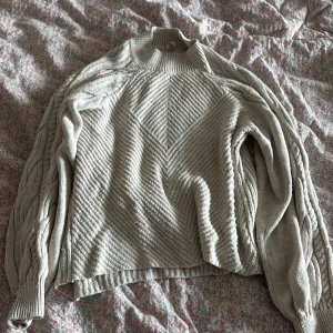 Beige stickad tröja med flätmönster - Säljer en beige stickad tröja med snyggt ribbat mönster framtill och flätade detaljer längs ärmarna. Tröjan har en hög krage och långa ärmar, perfekt för kyliga dagar. Passar dig som gillar en chill och stilren look.