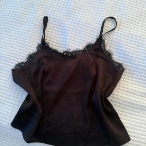 Svart linnetopp med spetskant - Snygg svart linnetopp med tunna axelband och dekorativ spetskant längs halsringningen. Toppen har en mjuk, glansig yta som ger en lyxig känsla och är perfekt för att lyfta din outfit. Passar dig som gillar stilrena och trendiga plagg.