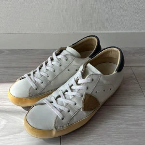 Philippe Model  - Snygga vita sneakers från Philippe Model med beige mockadetaljer på sidorna och bak, samt svart läderpanel vid hälen. Skorna har vit snörning och en rund tå, med en krämfärgad platt sula. Perfekt för dig som gillar stilrena och klassiska sneakers med en twist. Strl 39