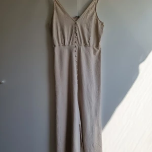 Ljus beige linneklänning Zara - Säljer en stilren beige långklänning från Zara med v-ringad hals och smala axelband. Klänningen har dekorativa knappar framtill och är tillverkad i linne och viskos. Passformen är normal och modellen är ärmlös, perfekt för varma dagar.