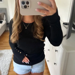 Svart långärmad stickad tröja med knappar - Svart stickad tröja med rund hals och långa ärmar. Snygga guldfärgade knappar på ena ärmen ger en extra detalj. Passformen är normal och materialet känns mjukt och skönt mot huden. Perfekt att matcha med jeans eller shorts för en chill look.