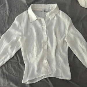 Vit skjorta i silkeslook från NA-KD - Snygg vit skjorta från NA-KD i storlek 36. Skjortan har en silkeslen finish, klassisk krage och knappar framtill. Långärmad och lätt transparent, perfekt för en clean och fräsch stil. Passformen är normal och materialet känns lyxigt. Den är även lite figursydd
