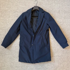 Mörkblå rock från Canali - Snygg mörkblå rock från italienska Canali, tillverkad i vattenavvisande material. Klassisk design med krage, två knappar framtill och praktiska sidfickor. Perfekt för dig som vill ha en stilren och exklusiv look under höst oh vinter. Köpt på NK för 12 000kr. Passar storlek M-L