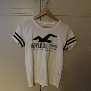Vit Hollister t-shirt med svarta ränder - Vit t-shirt från Hollister med svart logga och text framtill. Svarta dubbla ränder på ärmarna ger en sportig vibe. Rund hals och slim fit passform. Tillverkad i mjuk bomull som är skön mot huden. Perfekt för chill dagar och häng med kompisar.