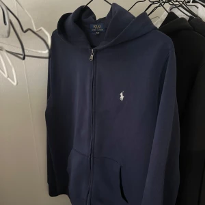 Mörkblå hoodie från Polo Ralph Lauren - Säljer en mörkblå hoodie från Polo Ralph Lauren med dragkedja och klassisk vit logga på bröstet i 10/10 skick. Tröjan har huva, fickor framtill och är tillverkad i mjuk bomull. Pris kan diskuteras vid snabb affär