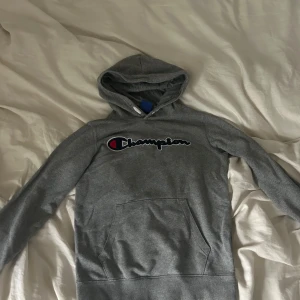 Grå hoodie från Champion - Säljer en grå hoodie från Champion med stor logga broderad på bröstet. Tröjan har huva, magficka och långa ärmar. Perfekt för chill dagar och streetwear-stil. Materialet är mjuk bomull och polyesterblandning för extra komfort.