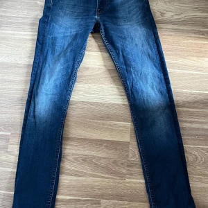 Slim fit blå jeans W29 L32 - Slim fit jeans från MVP i klassisk mörkblå tvätt med snygga slitningar framtill och bak. Modellen har raka ben och fem fickor, samt kontrastsömmar och knappstängning. Perfekt för dig som gillar en smal och modern siluett.