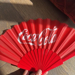 Röd Coca-Cola solfjäder - Cool röd solfjäder med klassisk Coca-Cola-logga i vitt. Gjord i lätt syntetmaterial och plast, perfekt att ta med till stranden eller festivalen. Fällbar design som är enkel att ha i väskan. Snygg accessoar för dig som vill sticka ut och hålla dig sval.