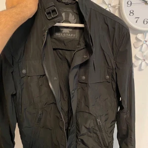 Svart vindjacka från Belstaff - Svart vindjacka från Belstaff med coolt quiltade paneler på sidorna och ryggen. Jackan har flera fickor med dragkedja och tryckknappar, samt en snygg ståkrage med spänne. Materialet är lätt och polyester, perfekt för blåsiga dagar. Belstaff-logga på ärmen.   