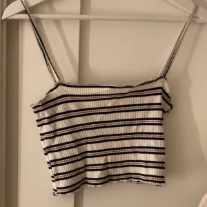 Randig ribbad linnetopp - Snygg croppad linnetopp med smala axelband och ribbad struktur. Toppen är vit med marinblå och beige ränder, perfekt för varma dagar. Enkel och stilren design som passar till jeans eller kjol.