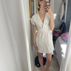 Vit omlottklänning med volangärm - Säljer en söt vit kortklänning med omlottmodell och knyt i midjan. Klänningen har volangdetaljer på ärmarna och en djup v-ringning. Perfekt för sommarens alla tillfällen, superfräsch och lätt att styla med sneakers eller sandaler.