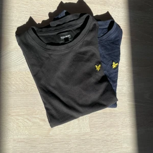Svart och marinblå t-shirt Lyle & Scott - Två stilrena t-shirts från Lyle & Scott Junior, en i svart och en i marinblå. Båda har rund hals och korta ärmar, med den klassiska gula fågel-loggan broderad på bröstet. 