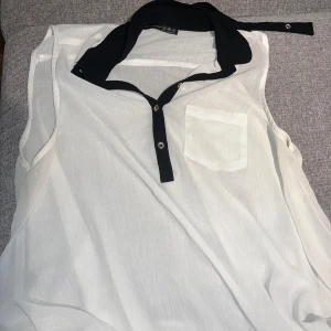 Vit ärmlös henley från Atmosphere - Snygg vit henley-skjorta från Atmosphere med svart krage och knappslå. Modellen är ärmlös och har en liten bröstficka. Materialet är lätt och luftigt, perfekt för varma dagar. Passar dig som gillar stilrena och enkla plagg med en twist.