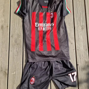 AC Milan fotbollströja R. Leão #17 - Säljer en AC Milan matchkit med R. Leão och nummer 17 på ryggen. Tröjan är svart med röda vertikala ränder, korta ärmar med italienska flaggans färger på mudden och broderat klubbmärke på bröstet. Materialet är lätt och andas, perfekt för fotboll.