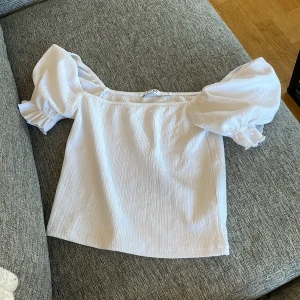 Vit blus med puffärmar från NA-KD - Supersöt vit blus från NA-KD med puffiga offshoulder-ärmar och volangdetaljer vid ärmslut. Materialet är lätt strukturerat och känns luftigt, perfekt för varma dagar. Blusen har en loose passform och ger en romantisk vibe till din outfit.