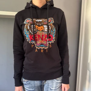 Svart Kenzo hoodie  - Svart hoodie från Kenzo med ikoniskt broderat tigermotiv och färgglada detaljer på bröstet. Tröjan har huva med snörning, ribbade muddar och är tillverkad i 100% bomull. Passformen är normal och den har långa ärmar. Perfekt statement-plagg för dig som gillar streetstyle.