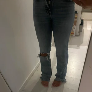 Botcut jeans Gina - Säljer ett par blå jeans som är midwaist. Jeansen har ett snyggt hål med slitning på ena knät och råa kanter nertill. Säljer då dom är för små.