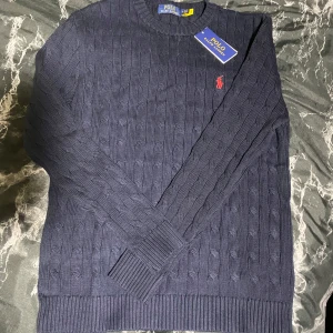 Marinblå stickad tröja Polo Ralph Lauren - Snygg marinblå stickad tröja från Polo Ralph Lauren med klassiskt kabelstickat mönster och röd broderad logga på bröstet. Rund hals och långa ärmar med ribbade muddar. Perfekt för dig som gillar stilrena och tidlösa plagg. Skriv gärna om mer bilder eller frågor