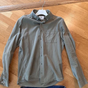 Grön skjorta från Ciszere med knappar - Snygg grön skjorta från Ciszere med klassisk button-down krage och bröstficka. Skjortan har långa ärmar och är tillverkad i mjuk bomull som känns skön mot huden. Perfekt för dig som gillar stilrena och enkla plagg med en chill vibe.