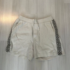 Beiga shorts med mönstrade sidor H&M - Snygga beiga shorts från H&M med relaxed fit och svartvit mönstrad kant längs sidorna. De har snörning i midjan och är tillverkade i mjuk bomull, perfekta för chill dagar eller när du vill ha en avslappnad stil.