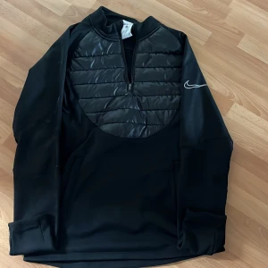 Svart Nike half zip tröja - Svart långärmad tröja från Nike med quiltad frontpanel och half zip-dragkedja. Swoosh-logga på ärmen ger en sportig vibe. Materialet är polyester och syntet, perfekt för träning eller chill. Snygg och funktionell design som sticker ut.
