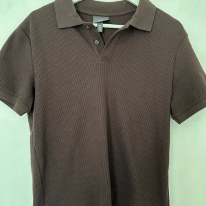 Brun ribbad slim fit t-shirt H&M - Snygg brun t-shirt från H&M med ribbad struktur och klassisk krage. Slim fit-modell med korta ärmar och två knappar vid halsen. Perfekt för dig som gillar stilrena och enkla plagg med lite edge.