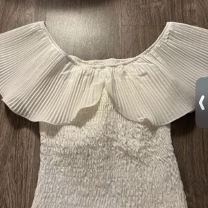 Vit offshoulder kortklänning - Supersöt vit kortklänning med offshoulder och bred plisserad volang upptill. Smockad överdel ger en skön och figurnära passform. Perfekt för sommarens alla tillfällen, enkel att styla med smycken eller sneakers.