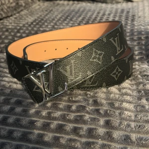 Louis Vuitton svart skinnbälte - Snyggt svart bälte från Louis Vuitton med klassiskt monogrammönster och stor LV-spänne i silver. Bältet är gjort i exklusivt skinn med beige insida och har en bred, stilren form som verkligen sticker ut. Perfekt för dig som gillar lyxiga accessoarer.