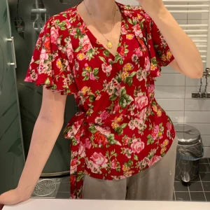 Röd omlottblus med blommigt mönster - Säljer en röd omlottblus med blommigt mönster i rosa, grönt och gult. Blusen har volangiga korta ärmar och knyt i midjan för en snygg siluett. V-ringad hals och mjukt, lätt material som känns skönt mot huden. Perfekt för dig som gillar färg och mönster! Helt oanvänd.