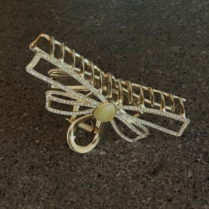 Hårklämma  - Superfin hårklämma i guld med en stor rosettformad design. Klämman är dekorerad med små glittriga stenar längs kanterna och har en pärlliknande detalj i mitten. 