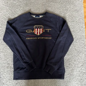 Mörkblå sweatshirt från GANT - Snygg mörkblå sweatshirt från GANT med broderad logga och sköld på bröstet. Tröjan har rund hals, långa ärmar och ribbade muddar. Storleken är 158-164, 13-14 år. Köpt på kids brand store och använd 3 gånger.