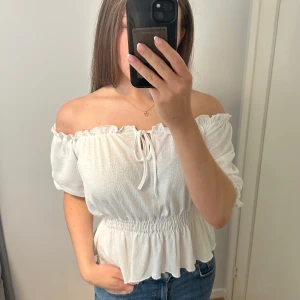 Vit offshoulder blus med volang - Superfin vit offshoulder blus med smockad midja och volangkant. Toppen har korta puffärmar och knytning framtill, liten sminkfläck på ena snöret (sista bilden) men inget man tänker på. Använd fåtal gånger💞