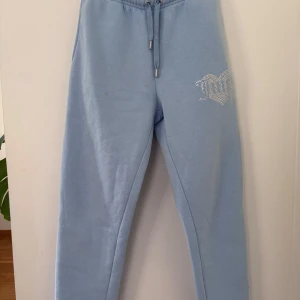 Blå mjukisbyxor Juicy Couture S - Snygga ljusblå mjukisbyxor från Juicy Couture med glittrande strassdetalj i form av ett hjärta och texten 'Juicy' på benet. Byxorna har snörning i midjan och är gjorda i mjukt bomullsmaterial. Perfekta för chill eller häng med kompisar.