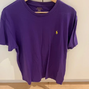 Lila t-shirt från Polo Ralph Lauren - Snygg lila t-shirt från Polo Ralph Lauren i storlek M. Klassisk design med rund hals och korta ärmar. På bröstet finns det ikoniska gula Polo-logot broderad. T-shirten är gjord i mjuk bomull och har en normal passform.