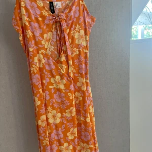 Blommig orange klänning från H&M Divided - Säljer en färgstark kortklänning från H&M Divided i orange med stora blommor i ljusgult och lila. Klänningen har omlottknytning fram och är ärmlös, perfekt för varma dagar. Materialet är mjukt och lätt, troligen polyester. Supersnygg till sneakers eller sandaler!