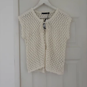 Stickad väst med knyt i offwhite - Supersöt stickad väst från Minou i offwhite med fint hålmönster och knyt framtill. Västen har vågiga kanter och är perfekt att slänga över en t-shirt eller klänning för en chill och trendig look.