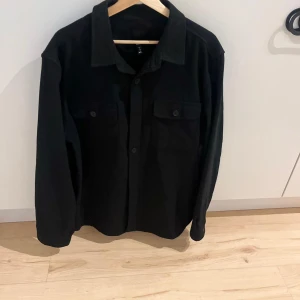 Svart overshirt från H&M XL - Stilren svart overshirt från H&M i regular fit. Skjortan har klassisk krage, två bröstfickor med knappar och långa ärmar. Perfekt att slänga över en t-shirt för en clean look. Materialet känns mjukt och passar bra till lager-på-lager.