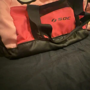 Rosa och röd träningsväska från SOC - Stor och rymlig träningsväska från SOC i rosa och rött med svarta detaljer. Väskan har breda handtag, dragkedja upptill och flera fack. Perfekt för gymmet eller sportaktiviteter. Tillverkad i slitstark polyester.