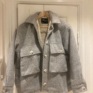 Grå overshirt jacka från Fanny Lyckman - Snygg grå overshirt jacka från Fanny Lyckman med stora bröstfickor och knappar framtill. Jackan har en mjuk ullkänsla och är fodrad med teddy på insidan för extra värme. Perfekt för lager-på-lager och chill höst/vinterstil.