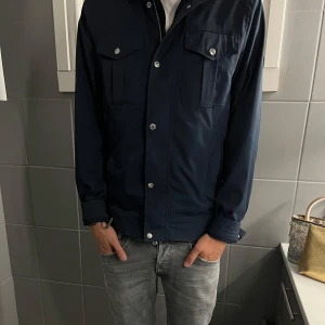 J.Lindberg Mörkblå Fieldjacket   - Vi säljer nu denna stilrena mörkblåa fältjavka i storlek S, skick 10/10. jackan är byggd med raka linjer och två bröstfickor med lock och tryckknappar. Jackan har hög krage, långa ärmar och silverfärgade knappar framtill. Jackan är perfekt till dig som letar efter plagg nu inför hösten/vintern. Pris kan diskuteras vid snabb affär och vid fler frågor är det bara att skriva!